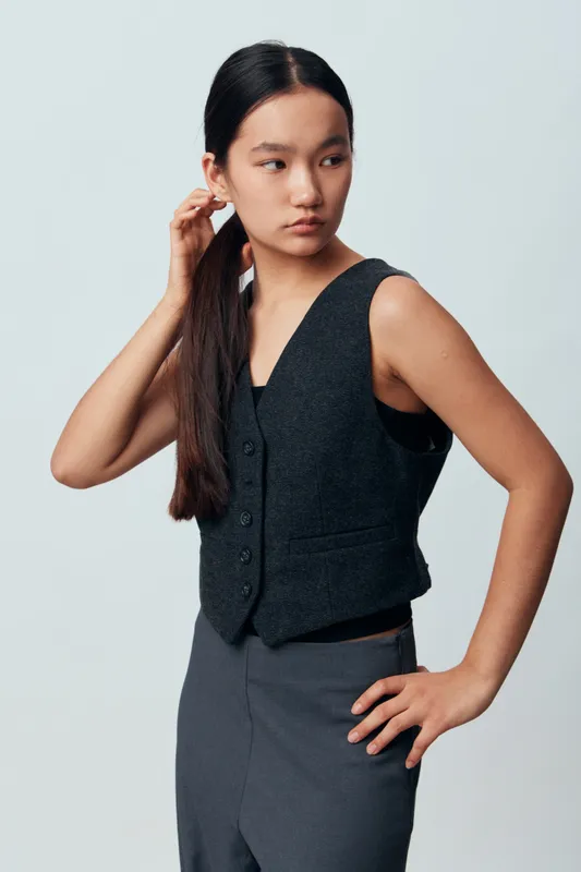 Young woman with long dark hair wearing black sleeveless vest and gray pants posing with hand on hip and looking sideways on light gray background in studio.