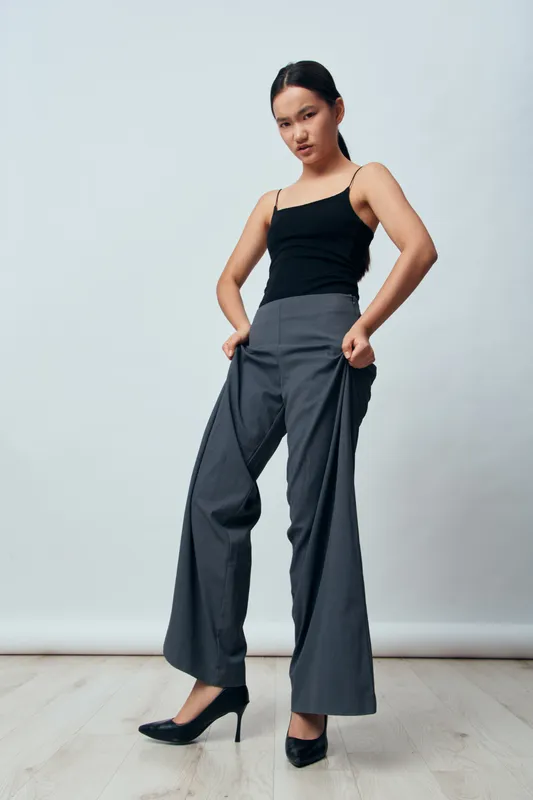 Woman wearing black top and gray wide leg pants standing with hands in pockets on wooden floor against white background in fashion studio portrait pose.