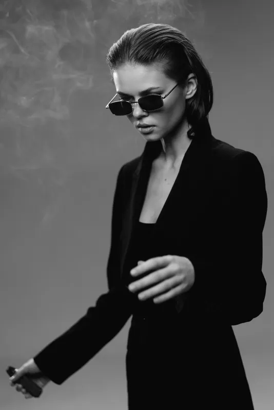 Stylish woman in sunglasses and black jacket with smoky background, confident pose, monochrome portrait, elegant fashion mood.