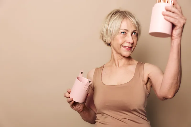 Mature woman with short blonde hair smiles while holding pink ceramic teapot and cup, wearing beige tank top against neutral background. Concept of tea time, relaxation, and lifestyle.