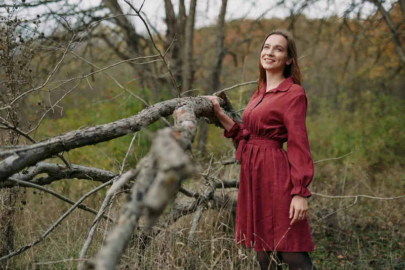A smiling woman in a red dress leans on a fallen tree in a quiet meadow, conveying an authentic, genuine connection to nature and a credible, everyday outdoor lifestyle moment.