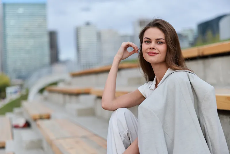 confident woman in casual light outfit sitting on urban outdoor bleachers with city background, natural smile and relaxed pose, daylight