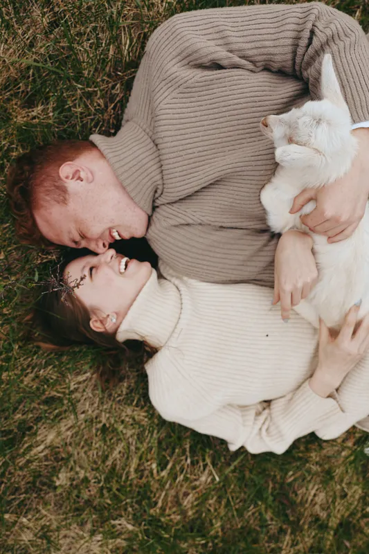 A couple lies on the grass smiling as a white goat kid rests on their chests, creating a warm countryside lifestyle moment with playful animal companionship outdoors.