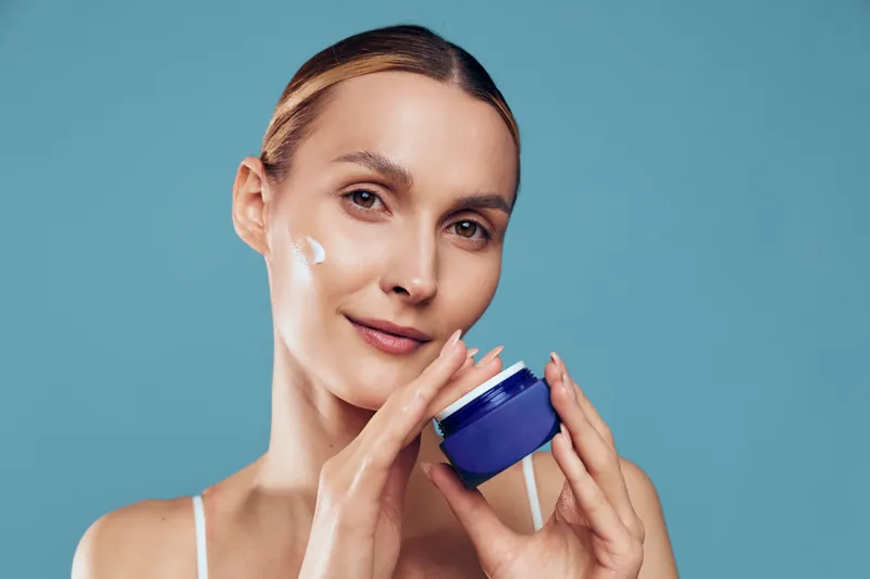 Skincare woman beauty face cream cosmetic moisturizer self care portrait, smiling with hand on cheek while holding blue jar, clean studio background, hydration concept