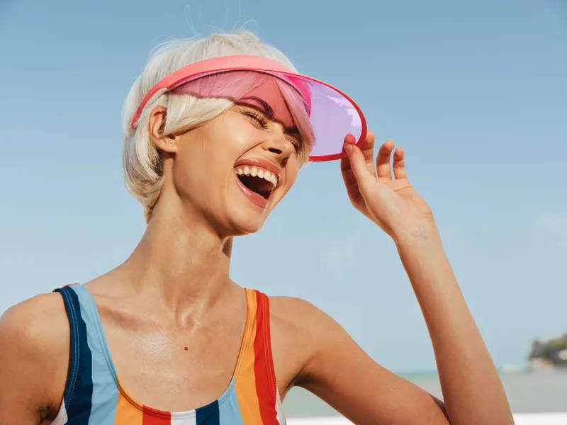 Energetic beach scene featuring a joyful woman wearing a colorful striped swimsuit and pink visor, laughing under a clear blue sky with spray and sunlit reflections.