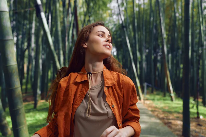 Woman forest bamboo portrait walking outdoor in orange jacket with gentle smile, looking up on a sunlit trail. Young traveler enjoying nature, peaceful green path and soft light.