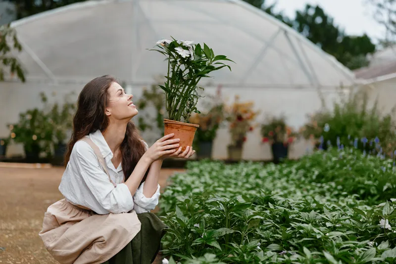 Young woman enjoying gardening in a greenhouse, holding a potted flower, surrounded by lush greenery and colorful plants, embodying joy and connection with nature