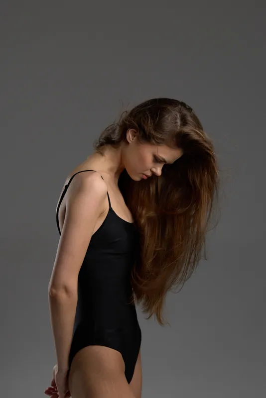 Woman with long hair in black bodysuit looking down, posing against gray background with soft lighting.