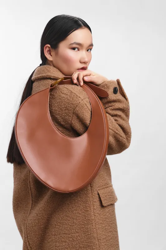 asian woman stands with a large brown bag over her shoulder in a minimalist studio, wearing a warm coat and calm expression, exuding chic confidence and poised street style.