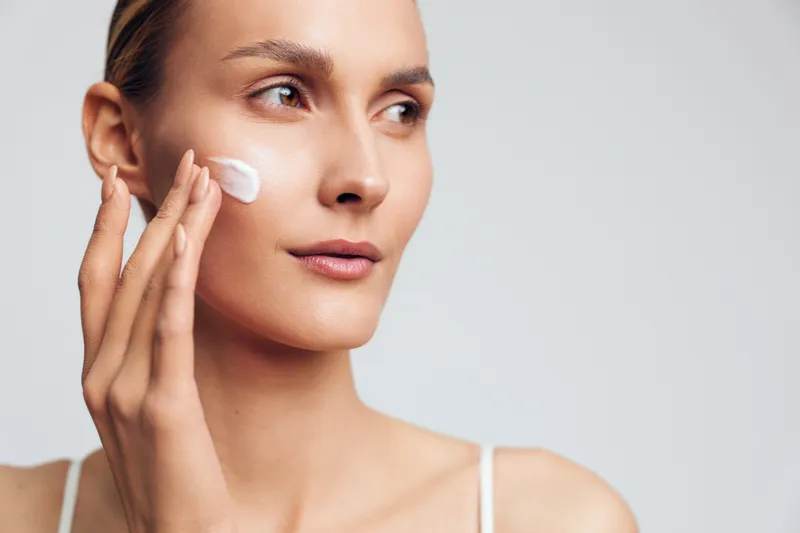 Young woman with clear skin applying white face cream on cheek, looking aside on light background. Concept of skincare, beauty routine, self-care, wellness, and natural complexion.