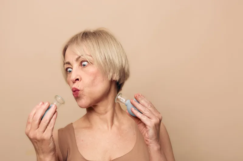 Middle aged woman with short blonde hair examines two small glass bottles with curious expression against beige background. Concept of curiosity, choice, and beauty product evaluation.