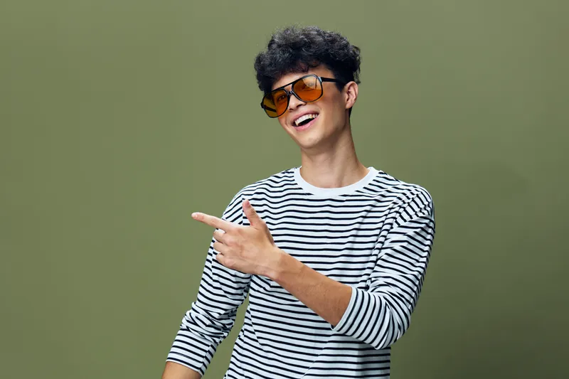 Smiling young man with curly hair wearing orange sunglasses and striped shirt pointing sideways against olive green background, casual style and joyful expression.