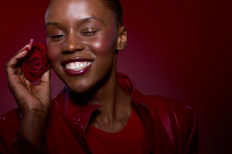 Close-up of smiling African American woman with dark skin holding red rose near face. She wears red leather jacket and red shirt on dark red background. Concept of beauty and happiness.