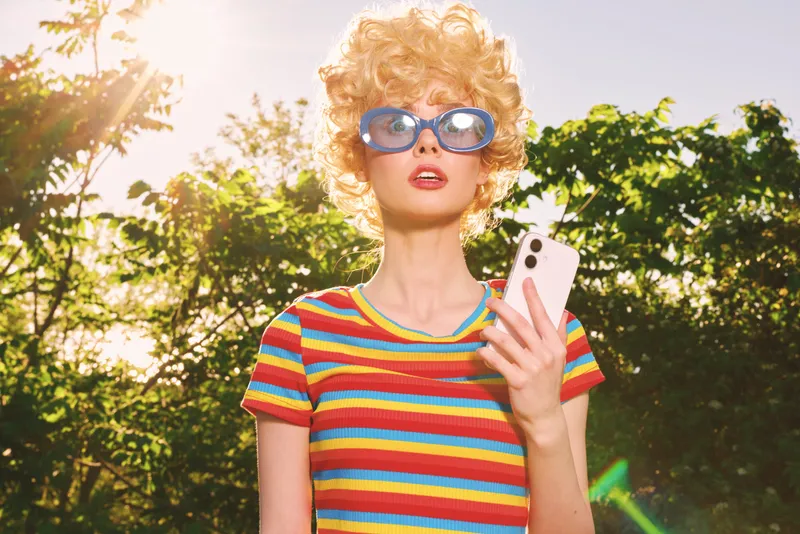 Smiling woman with curly hair wearing a colorful striped tee, holding a smartphone outdoors. Retro film color mood, lifestyle theme, sunny backdrop and warm, vibrant atmosphere.