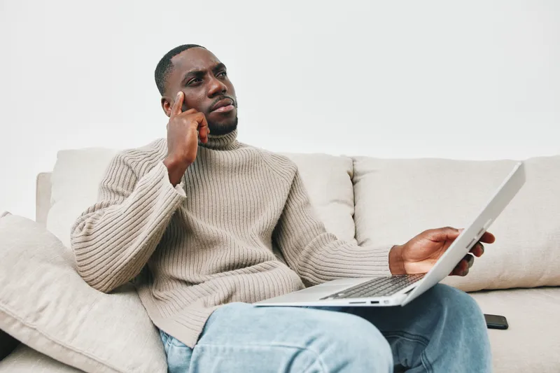Thoughtful Black man in a cozy sweater contemplating while working on a laptop, sitting on a comfortable couch in a bright, minimalistic room