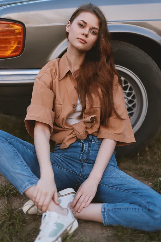 Young woman in casual fashion sitting by a vintage car, showcasing a relaxed style and natural beauty with a warm color palette.