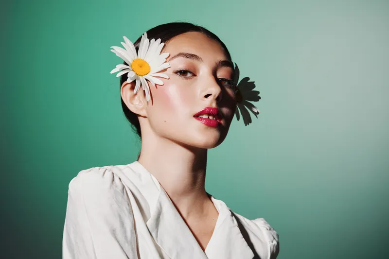 Elegant woman with dark hair wearing white blouse poses with daisies in her hair against teal background. Glamorous makeup and stylish concept for beauty, fashion, and floral creativity.
