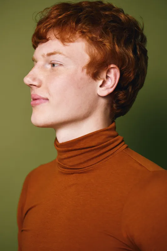 Young man with red hair wearing brown turtleneck sweater in profile, looking ahead with calm expression against green background in studio portrait.