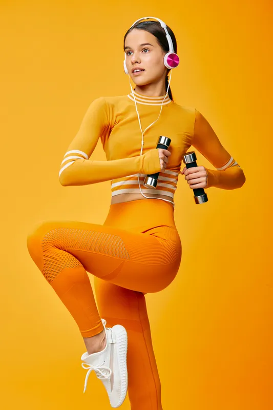 Woman fitness sport portrait in bright orange activewear, holding dumbbells and wearing headphones in a dynamic balance pose against a vivid monochrome studio background, energetic and focused.
