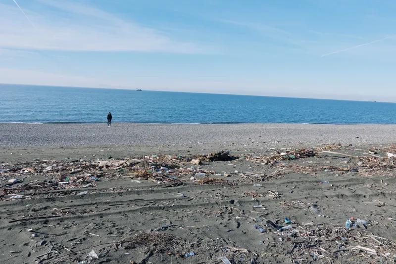 Deserted beach with litter along the shoreline, calm blue sea and clear sky, lone figure walking near trash on sand, highlighting pollution and environmental concerns