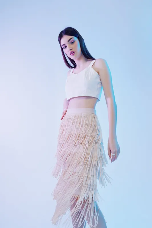 Young woman in a white crop top and fringe skirt poses against a light background. The image highlights beauty, fashion, and elegance with soft lighting and a modern style.
