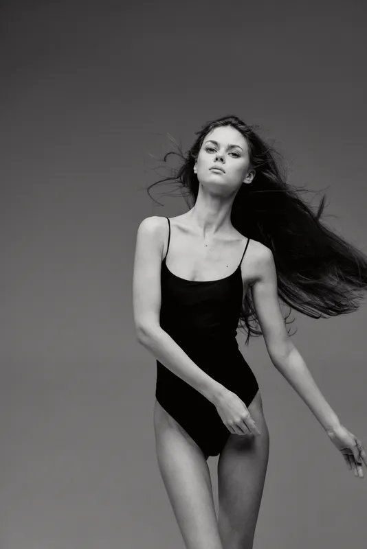 Black and white portrait of a slender woman in a black bodysuit with flowing hair and confident expression.