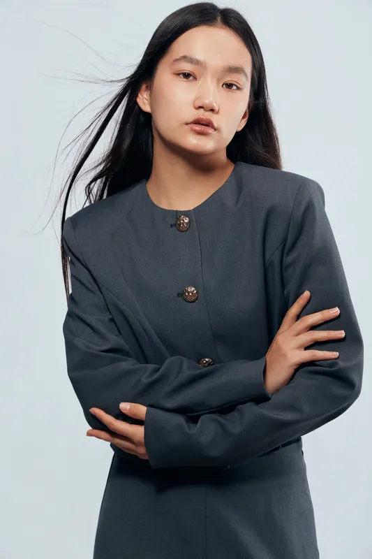 Young woman with long black hair wearing a gray dress with buttons poses against light background. Portrait of confident Asian female with crossed arms and calm facial expression.