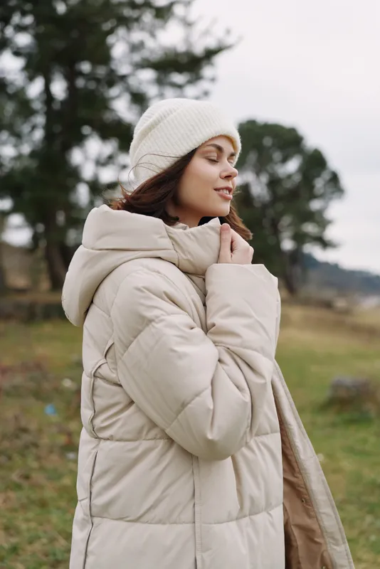 Winter woman outdoors in a beige coat, knit beanie and scarf, serene mood with soft natural light in an open field, conveying warmth, comfort and seasonal style.