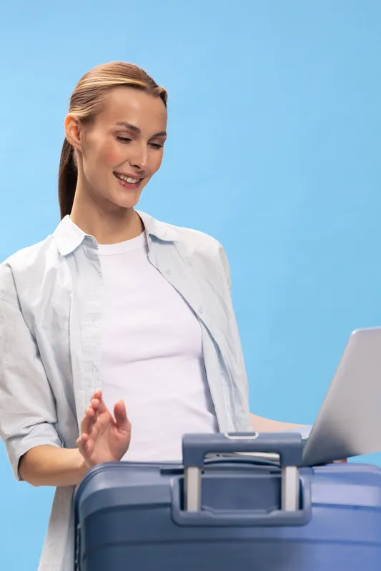 Young woman with ponytail standing near suitcase and laptop on blue background. She smiles and gestures with hand, concept for travel planning, vacation, and remote work balance.