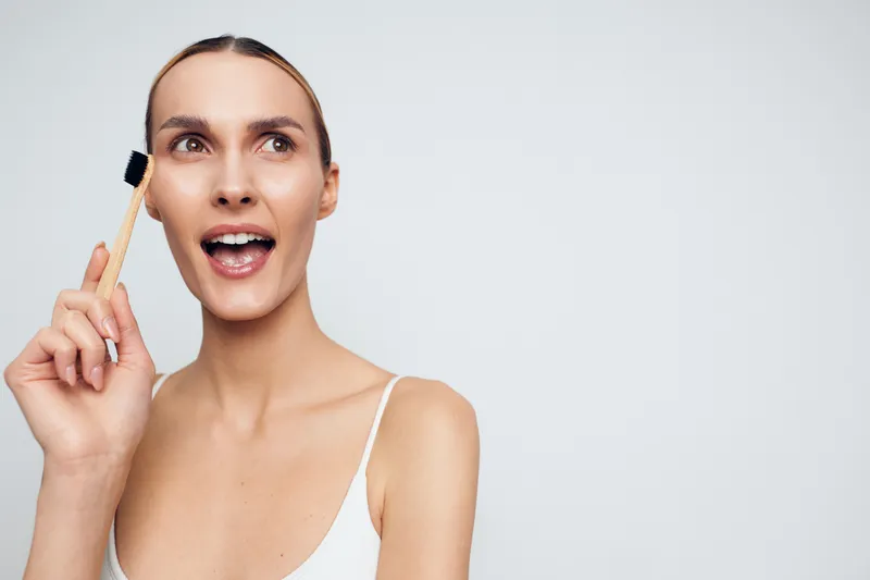 A young smiling woman in a white tank top holds a bamboo toothbrush near her face, posing against a plain white background. Concept for natural hygiene and eco friendly living.