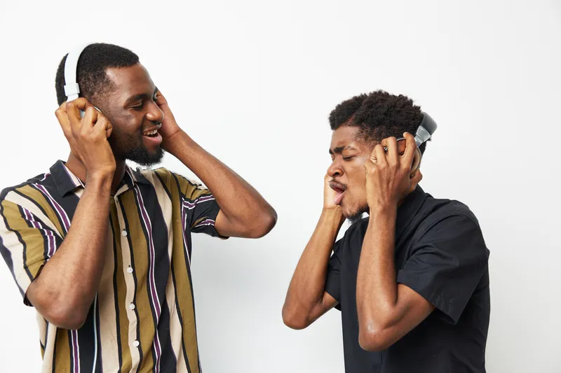 Two joyful black men enjoying music, wearing headphones and showcasing playful expressions against a light background. A vibrant and exciting atmosphere.