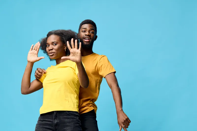 Young African couple smiling and gesturing with hands, wearing yellow outfits against a turquoise background, showcasing joy and connection