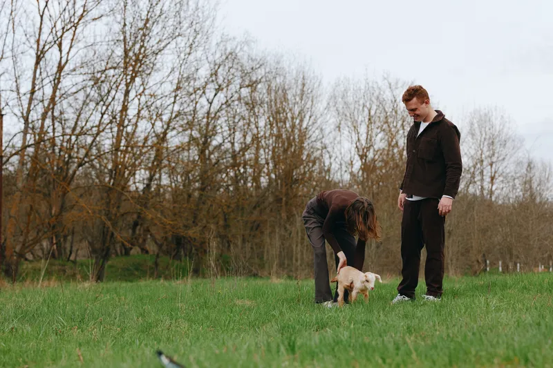 Casual lifestyle scene with two adults in casual clothes in a grassy field, bending to greet a small light dog, during an autumn afternoon in a tranquil park setting, with soft natural light leaves