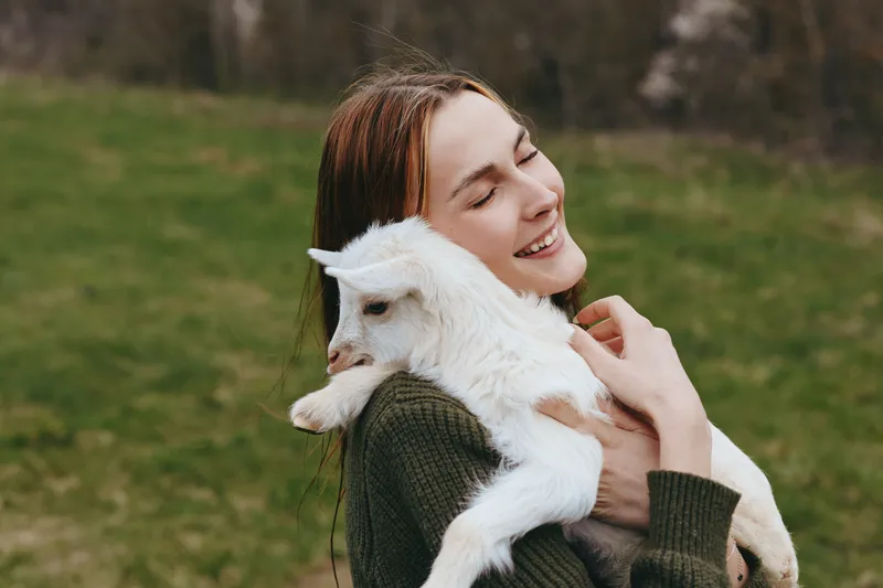 Cheerful woman outdoors, cuddling a goat kid in a sunlit meadow, relaxed smile, gentle breeze, natural colors, warm sweater, rustic charm, clear bond with animals, wholesome lifestyle moment