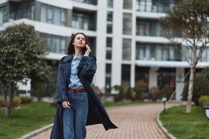 Young woman in a long dark coat walks confidently along a paved path near modern buildings while talking on her smartphone on a cloudy day.