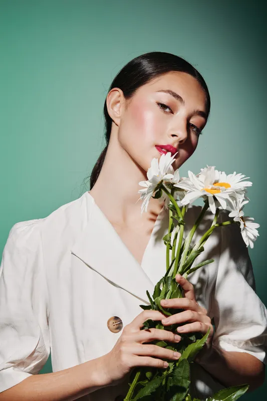 Young woman in white blouse holds white daisies close to face and looks at camera. Fresh beauty with fashionable makeup on pastel background. Concept of elegance and nature.