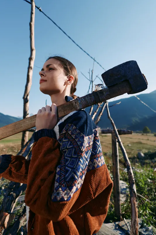 woman sledgehammer mallet rural vineyard mountains farmer outdoor carrying heavy wooden tool on shoulder, confident young agricultural worker in patterned jacket among trellis and fields under blue