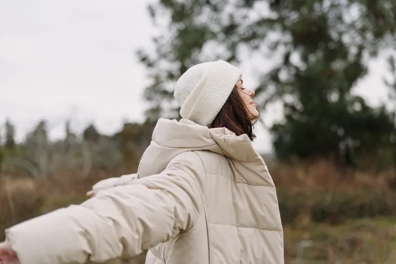 Woman outdoors in a beige winter coat and white beanie with arms outstretched, embracing fresh air and wide open space in a peaceful nature scene