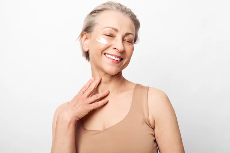 Smiling mature woman with short hair and facial cream patches on cheeks touches neck in beige tank top. Studio portrait on white background. Concept for skincare, beauty, and self care.