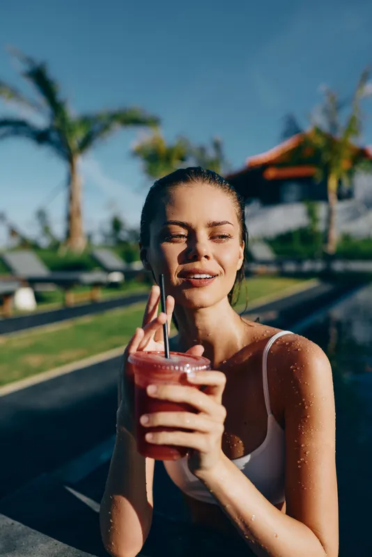 Woman by a sunny pool enjoying a refreshing smoothie, outdoors, smiling softly, casual summer vibe, palm trees in the background, warm light, candid lifestyle moment