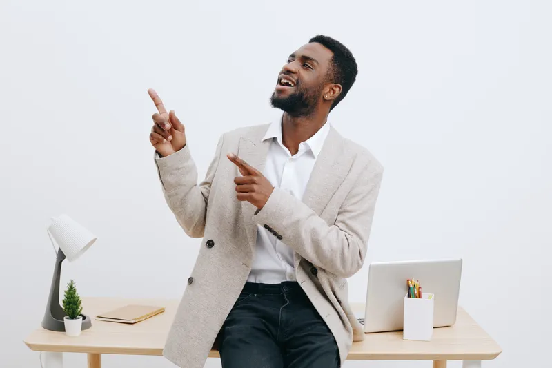 Smiling man with dark skin in casual beige blazer and white shirt standing in modern office space, pointing upward and looking happy. Business, teamwork, confident, professional, success, interior.