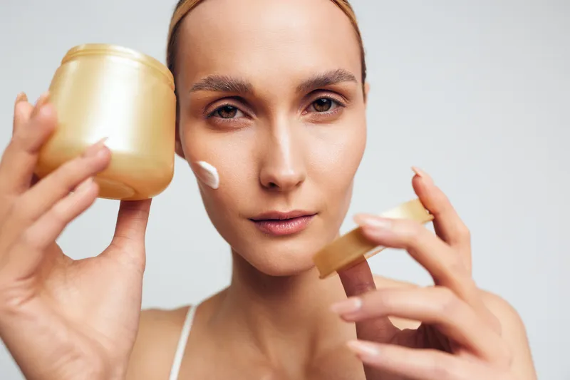 Young woman with clear skin applies cream on her face holding a gold jar and lid in a skincare routine against a light background with focused expression.