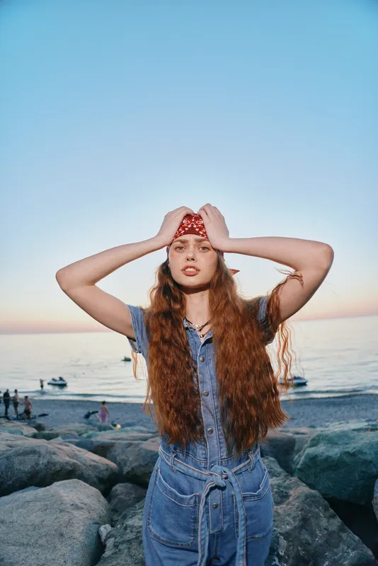 Lifestyle woman with long hair in modern boho-western denim street style stands by the rocky beach at sunset with film color giving a warm, vintage atmosphere.