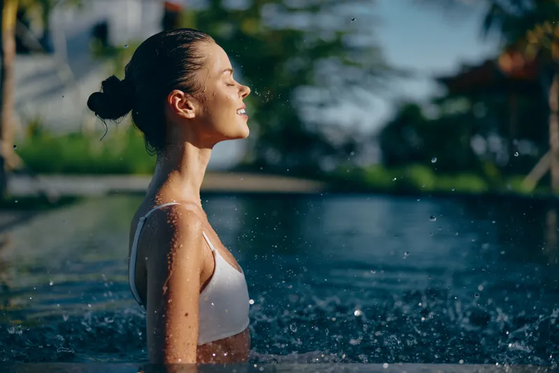 A woman stands in outdoor water, eyes closed, face tilted toward the sun as droplets spray around her. A moment of relaxation, freedom, and refreshing energy.