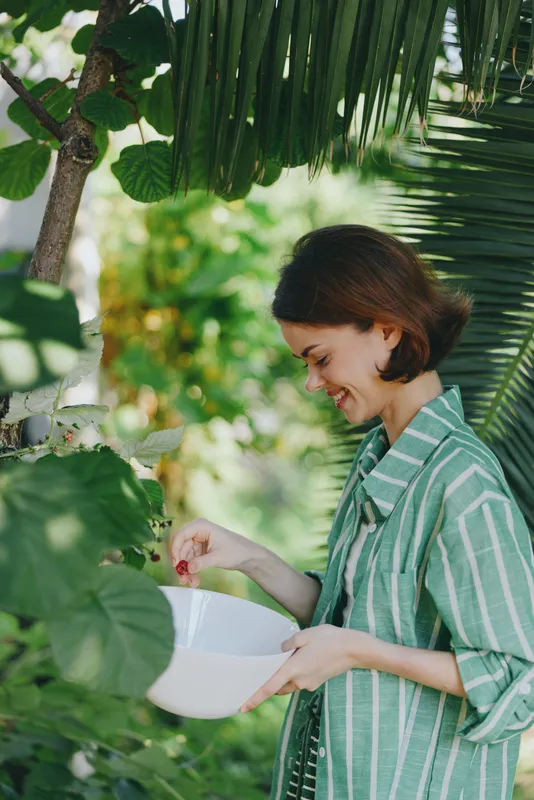 woman in a striped green shirt stands in a lush garden, holding a white bowl, smiling softly as natural light highlights her calm mood amid vibrant greenery