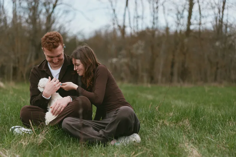 Couple sits on grass outdoors with a goat kid, sharing affection and gentle smiles while cradling the young animal in a candid lifestyle moment of bonding and nature