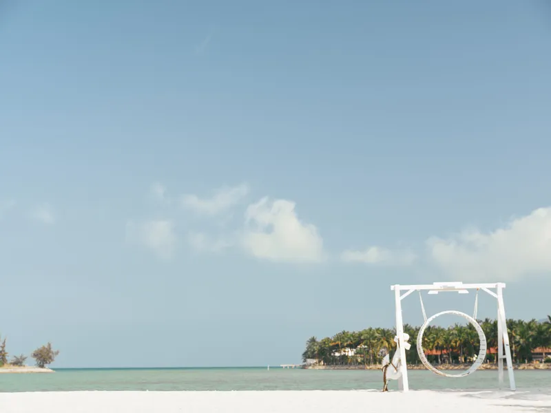 Tranquil beach scene featuring a circular swing on white sand, calm turquoise water, and a lone figure near the shore under a clear blue sky.