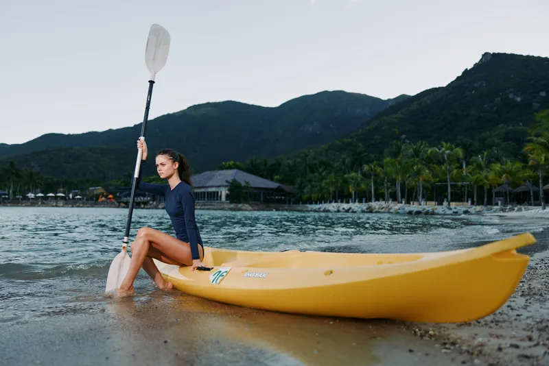 Woman sitting on yellow kayak at the shore holding paddle with mountains and palm trees in the background at sunrise.