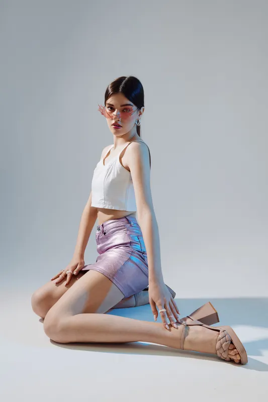 Young woman poses in a white crop top and shiny skirt with stylish sandals on a plain background. Concept of beauty, fashion, and modern style in studio lighting.