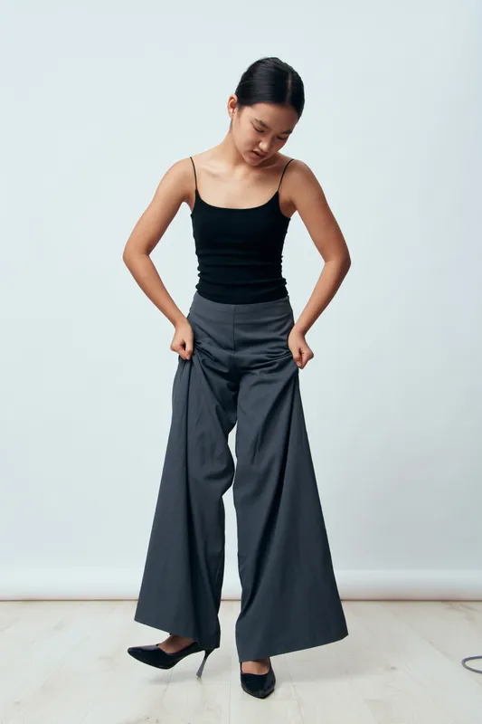 Young woman wearing wide leg gray pants and black sleeveless top stands on light floor against plain background looking down adjusting pants with black shoes on feet.